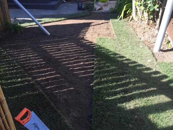 A garden bed part-laid with new turf over levelled soil, with a hand saw resting on the worked ground