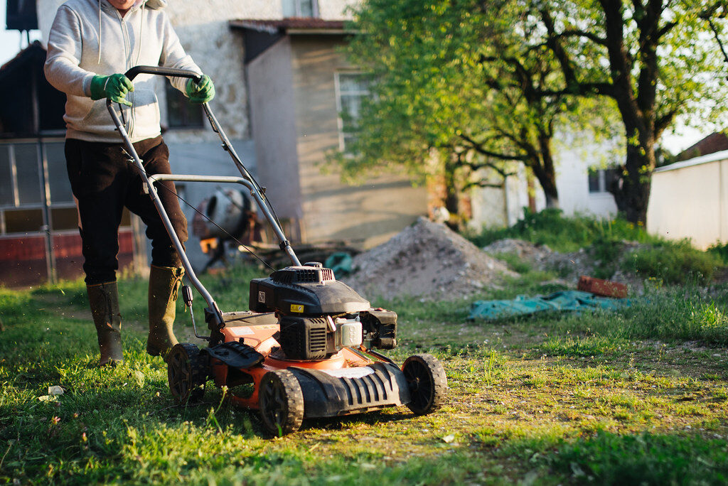 An operator in work clothes and gumboots pushing a petrol lawnmower across a residential backyard
